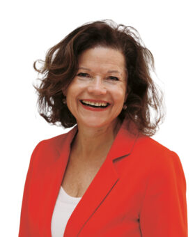 Eine Frau mit gewelltem braunem Haar, die einen leuchtend orangefarbenen Blazer und ein weißes Oberteil trägt, lächelt vor einem weißen Hintergrund.