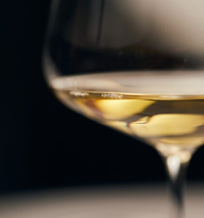 Nahaufnahme eines Weinglases, das teilweise mit Weißwein gefüllt ist, mit weicher Beleuchtung, die die goldene Farbe des Getränks hervorhebt, und einem verschwommenen dunklen Hintergrund.