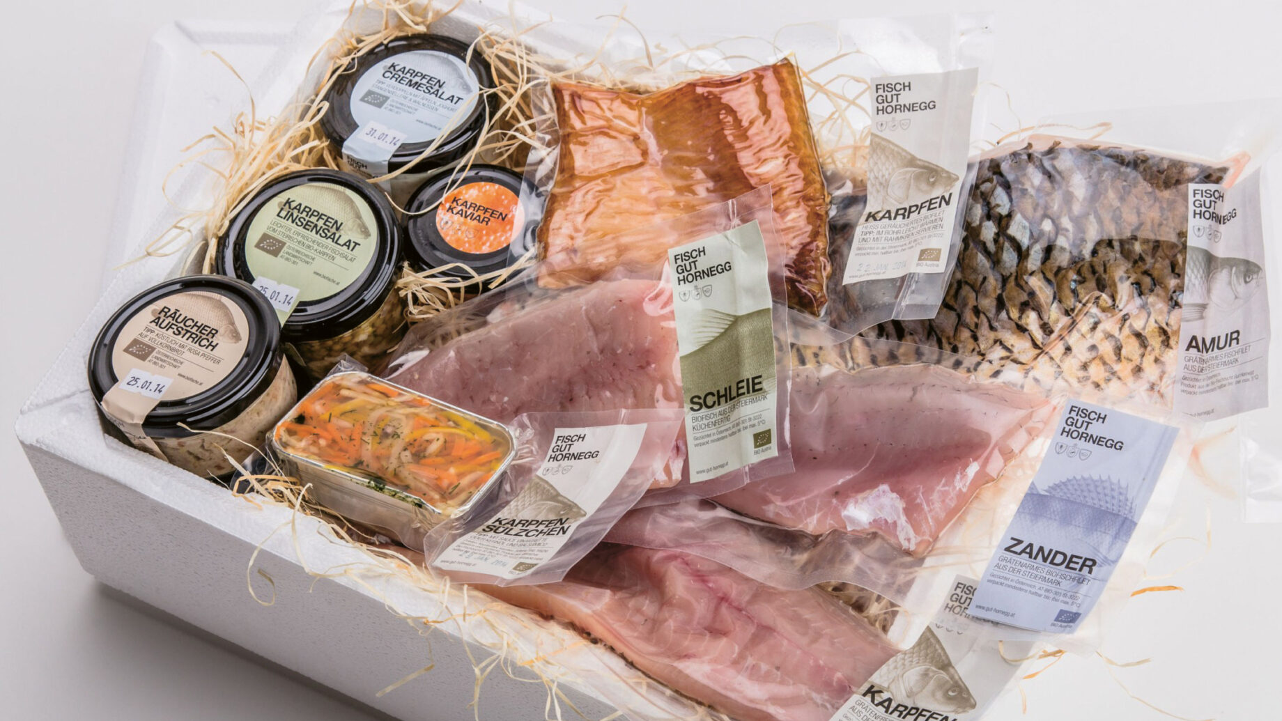 Sortierte verpackte Fischfilets und Aufstrichgläser in einer weißen Schachtel mit Strohfüllung, fertig für den Versand oder die Auslage.