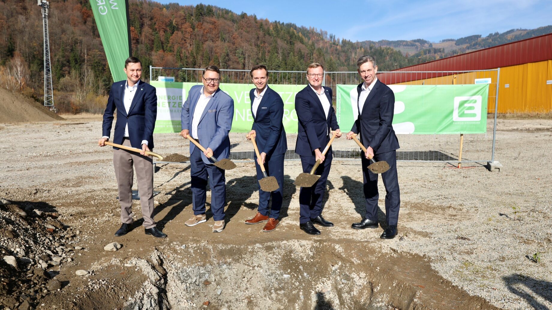 Fünf Männer in Anzügen stehen im Freien bei einer bahnbrechenden Veranstaltung als KS Engineers aus Graz treibt Elektromobilität an, mit Bannern und Hügeln hinter ihnen.