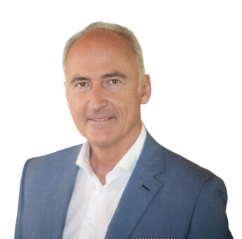 Lächelnder Mann mittleren Alters mit kurzen grauen Haaren in einem blauen Anzug und weißem Hemd, der vor einem schlichten weißen Hintergrund posiert.