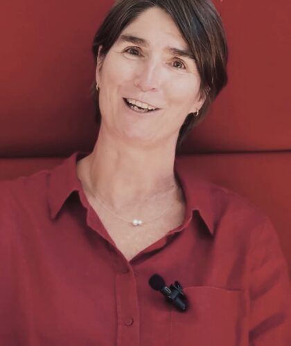 Eine Frau mit kurzen braunen Haaren, rotem Hemd und Brille auf dem Kopf, sitzt vor einem rot gepolsterten Hintergrund und lächelt. Sie hat ein kleines Mikrofon an ihr Hemd geklemmt.