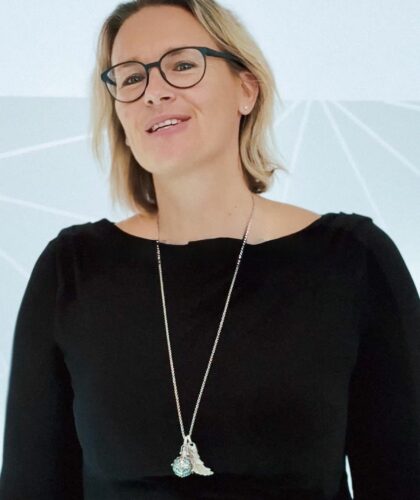 Eine Person, die eine Brille und ein schwarzes Kleid mit einer langen Halskette mit Anhänger trägt, steht vor einem hellen Hintergrund mit subtilen geometrischen Linien und lächelt leicht.