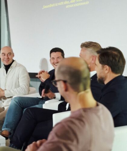 Fünf Männer sitzen bei einer Podiumsdiskussion auf der Bühne, einige lächeln und unterhalten sich.