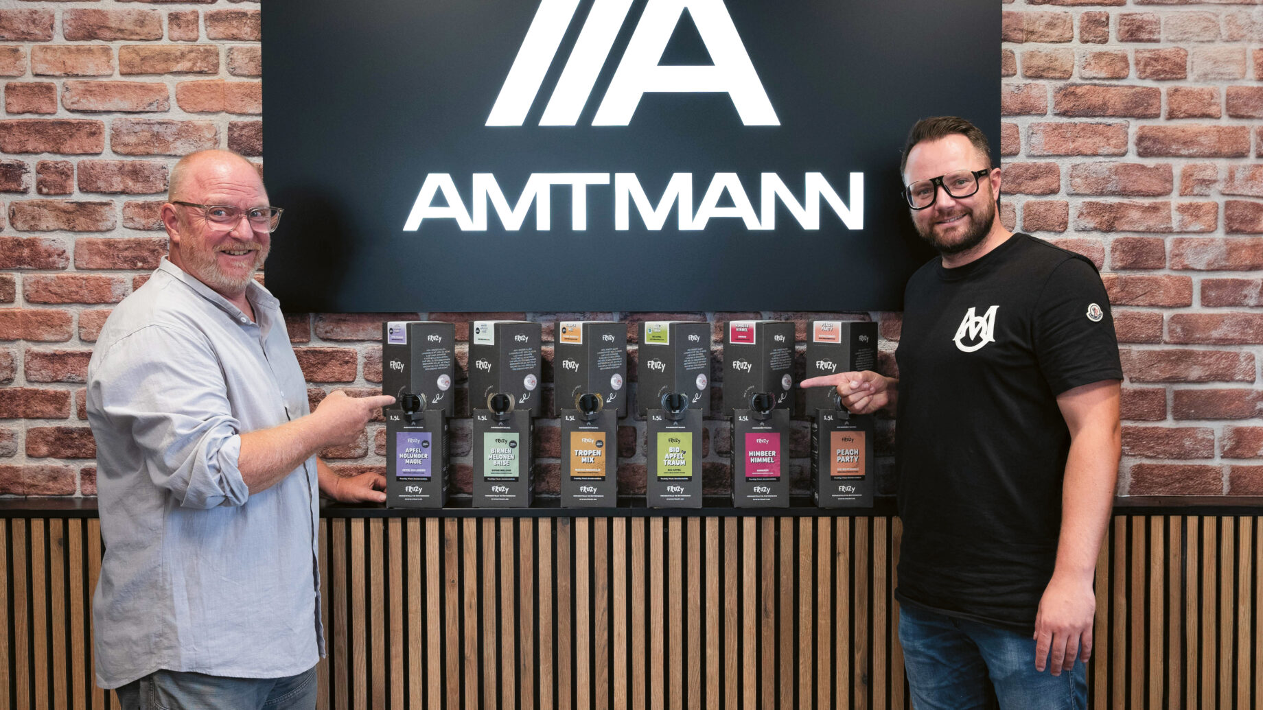 Zwei Männer stehen lächelnd vor einer Backsteinmauer und einem "AMTMANN"-Schild und zeigen jeweils auf eine Reihe verpackter Produkte, die auf einer Theke ausgestellt sind.