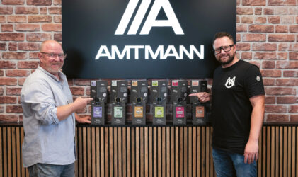 Zwei Männer stehen lächelnd vor einer Backsteinmauer und einem "AMTMANN"-Schild und zeigen jeweils auf eine Reihe verpackter Produkte, die auf einer Theke ausgestellt sind.
