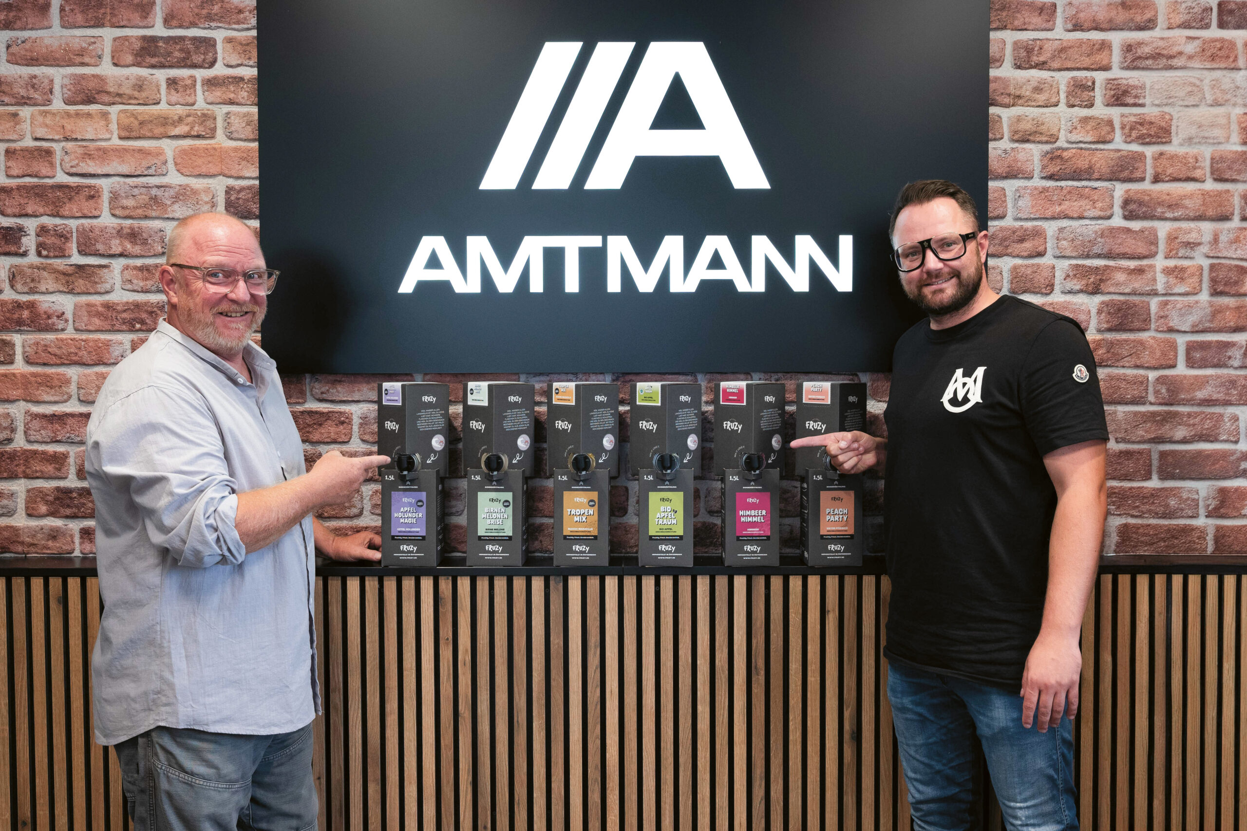 Zwei Männer stehen lächelnd vor einer Backsteinmauer und einem "AMTMANN"-Schild und zeigen jeweils auf eine Reihe verpackter Produkte, die auf einer Theke ausgestellt sind.