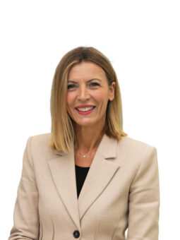 Eine Frau mit glattem blondem Haar trägt einen beigen Blazer und ein schwarzes Oberteil und lächelt vor einem schlichten weißen Hintergrund.