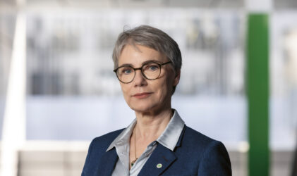 Eine selbstbewusste ältere Frau mit kurzen grauen Haaren und Brille, die einen blauen Blazer und eine silberne Bluse trägt, steht in einer modernen, hellen Büroumgebung.