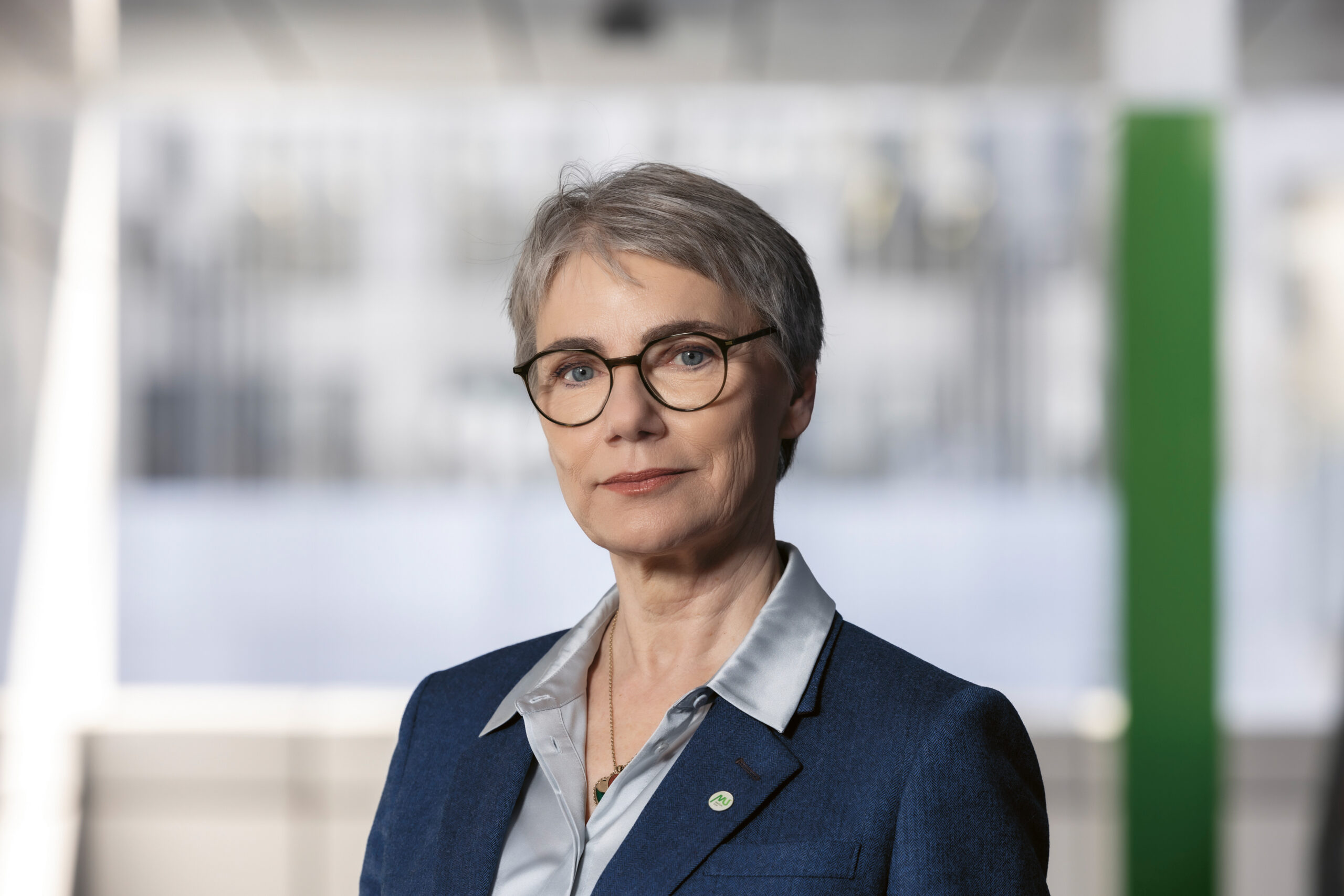 Eine selbstbewusste ältere Frau mit kurzen grauen Haaren und Brille, die einen blauen Blazer und eine silberne Bluse trägt, steht in einer modernen, hellen Büroumgebung.