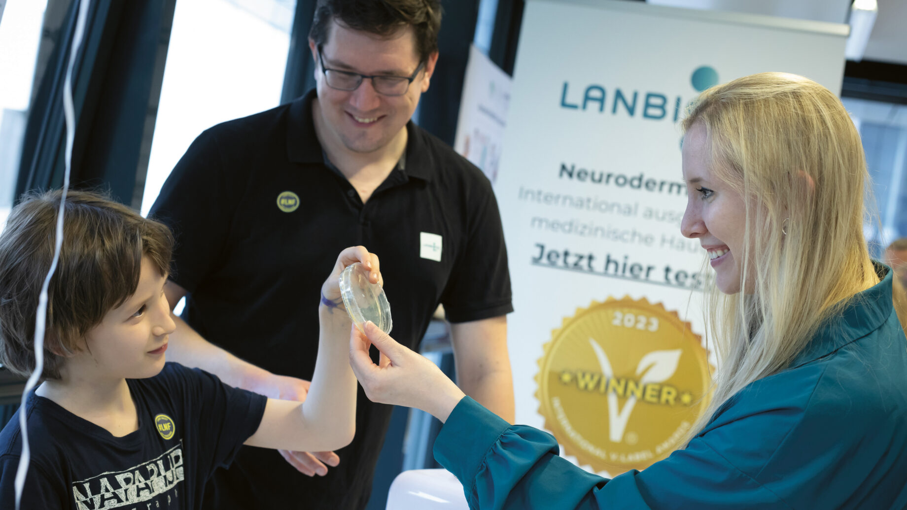 Eine Frau zeigt einem lächelnden Jungen ein durchsichtiges Objekt, während ein Mann zuschaut; sie stehen neben einem LANBIOTIC-Produktdisplay mit einer "Gewinner"-Plakette.