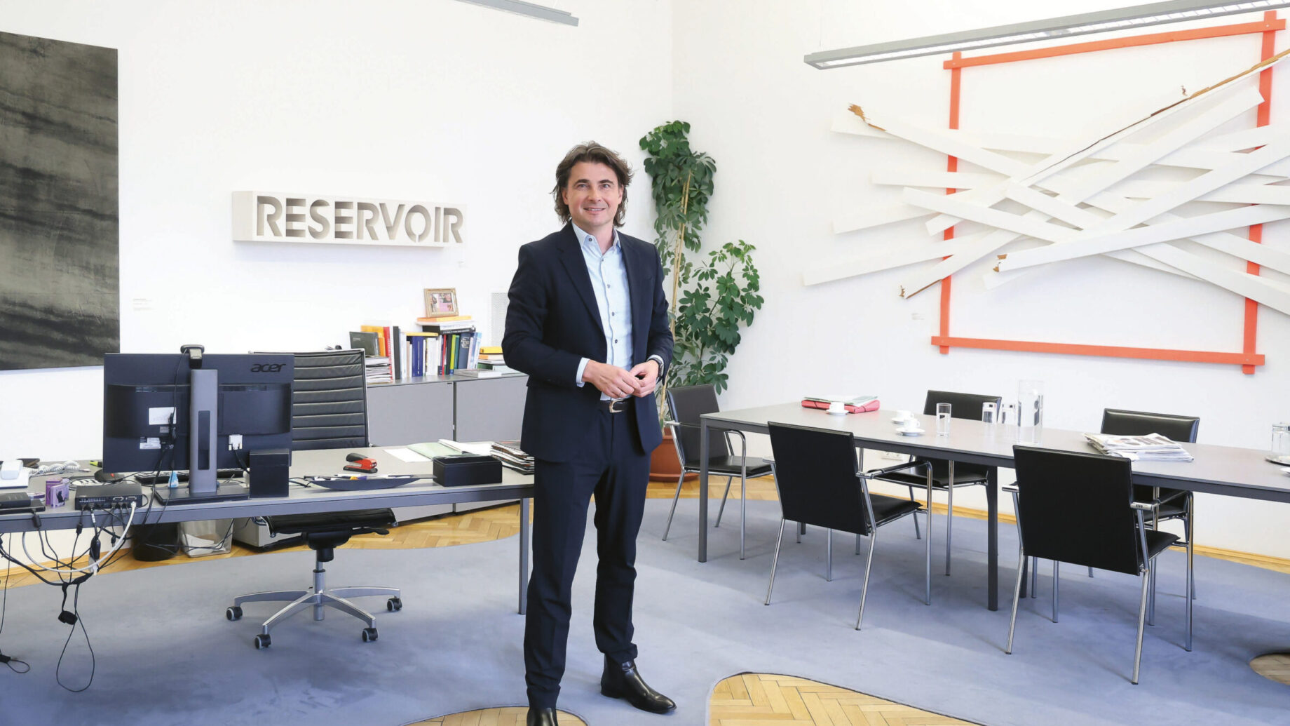 Ein Mann im Anzug steht lächelnd in einem modernen Büro mit Kunst an den Wänden, einem Schreibtisch mit Computermonitoren, einem Konferenztisch und Bücherregalen. Das Wort "RESERVOIR" steht an der Wand hinter ihm.