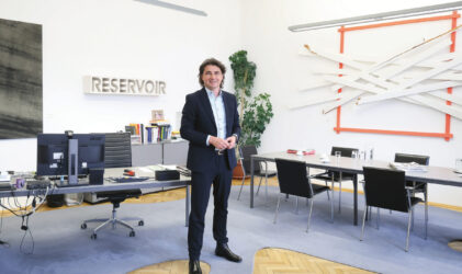 Ein Mann im Anzug steht lächelnd in einem modernen Büro mit Kunst an den Wänden, einem Schreibtisch mit Computermonitoren, einem Konferenztisch und Bücherregalen. Das Wort "RESERVOIR" steht an der Wand hinter ihm.