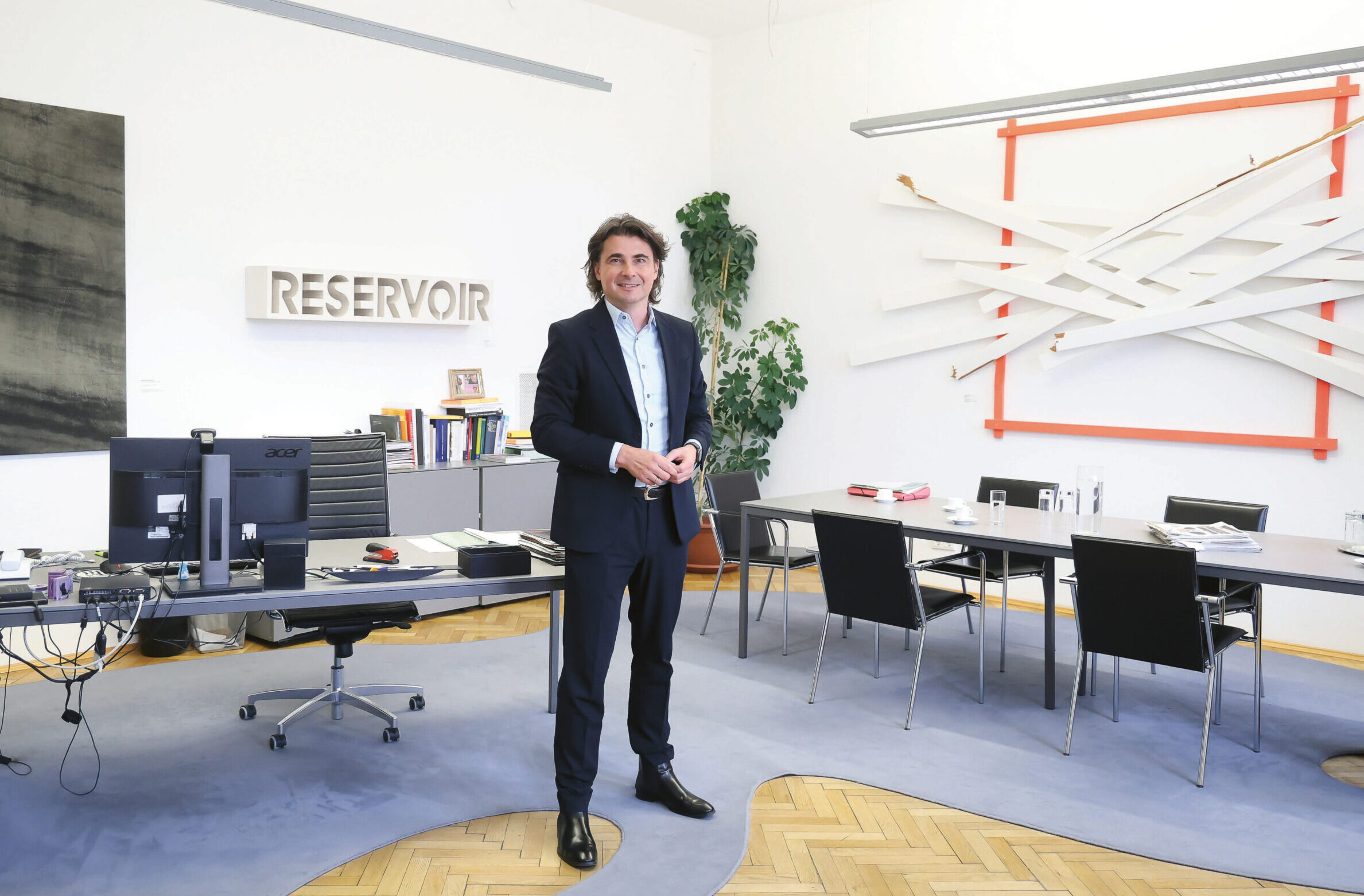 Ein Mann im Anzug steht lächelnd in einem modernen Büro mit Kunst an den Wänden, einem Schreibtisch mit Computermonitoren, einem Konferenztisch und Bücherregalen. Das Wort "RESERVOIR" steht an der Wand hinter ihm.