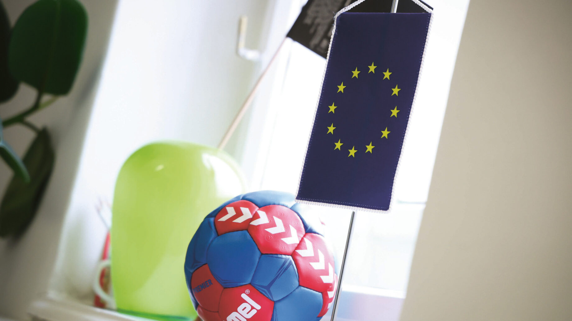 Eine kleine Flagge der Europäischen Union hängt neben einem bunten Handball und einem grünen Luftballon auf einer weißen Fensterbank.