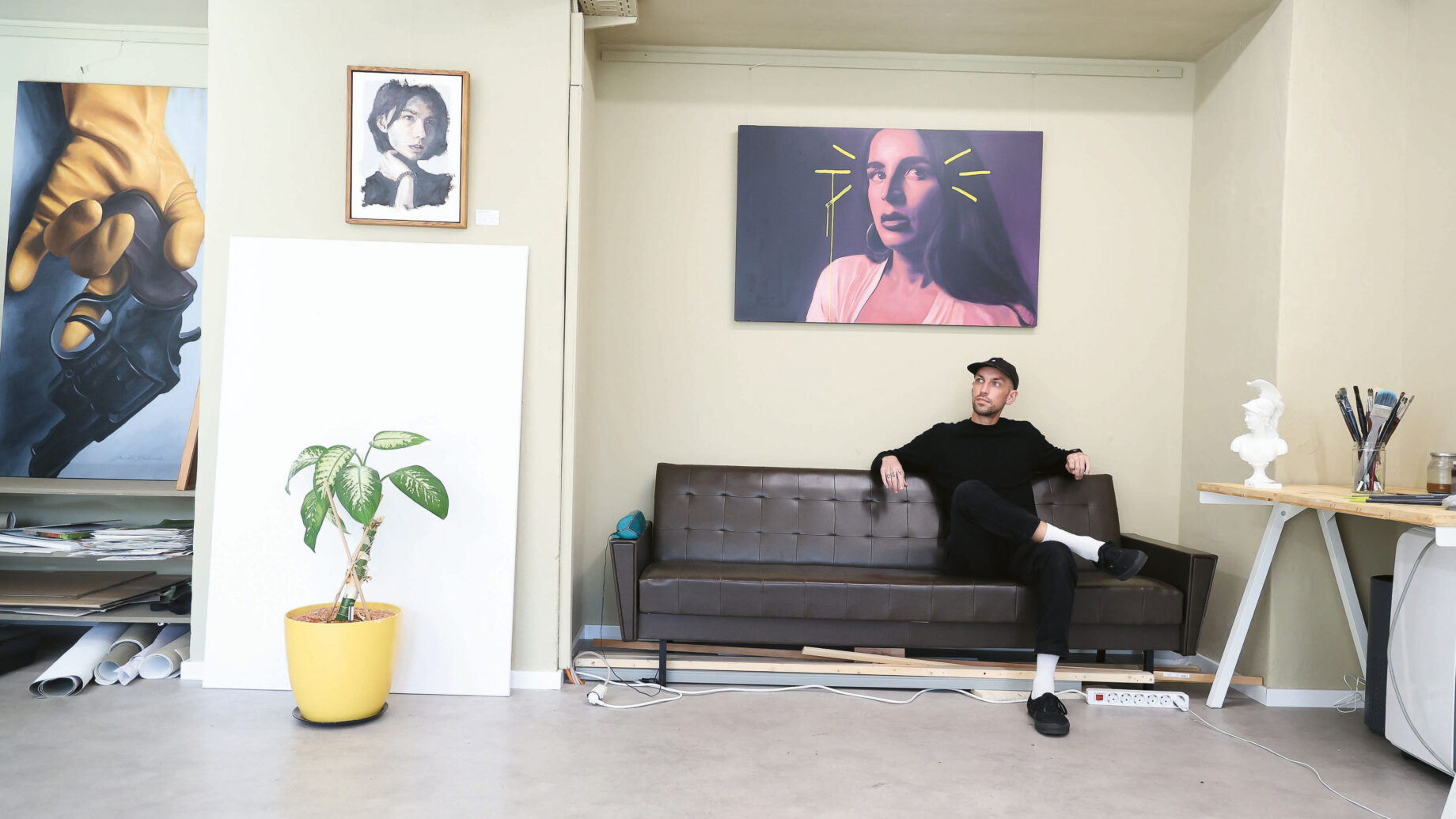 Ein Mann in schwarzer Kleidung und einer Mütze entspannt sich auf einem braunen Sofa in einem Kunstatelier, umgeben von Gemälden, einer gelben Topfpflanze, einem Tisch mit Malutensilien und dem Bild einer Frau, das über ihm an der Wand hängt.