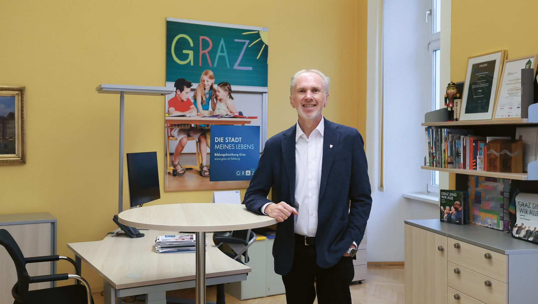 Ein Mann im Anzug steht lächelnd in einem hellen Büro mit gelben Wänden, Holzböden, Schreibtischen, Bücherregalen und einem Graz-Poster an der Wand.