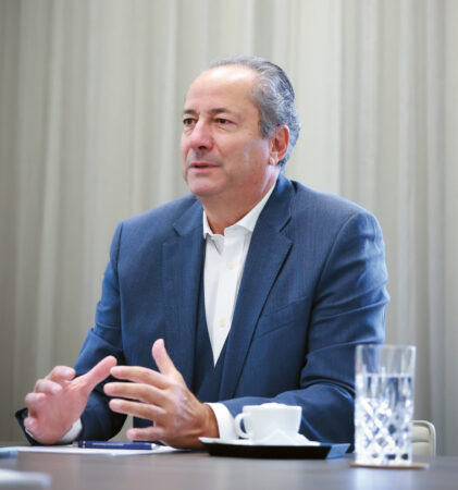 Ein Mann in einem blauen Anzug sitzt an einem Tisch mit einem Glas Wasser und einer Tasse und gestikuliert mit seinen Händen, während er spricht. Im Hintergrund sind helle Vorhänge zu sehen.