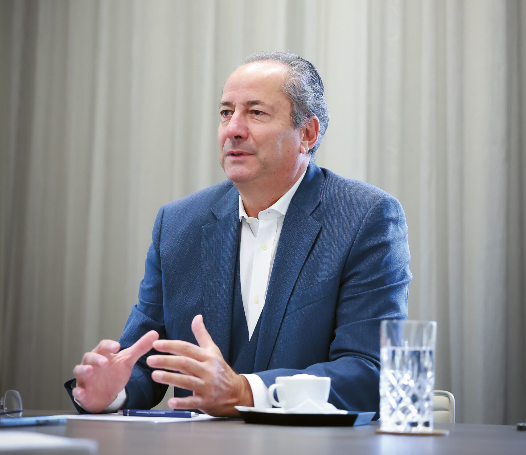 Ein Mann in einem blauen Anzug sitzt an einem Tisch mit einem Glas Wasser und einer Tasse und gestikuliert mit seinen Händen, während er spricht. Im Hintergrund sind helle Vorhänge zu sehen.