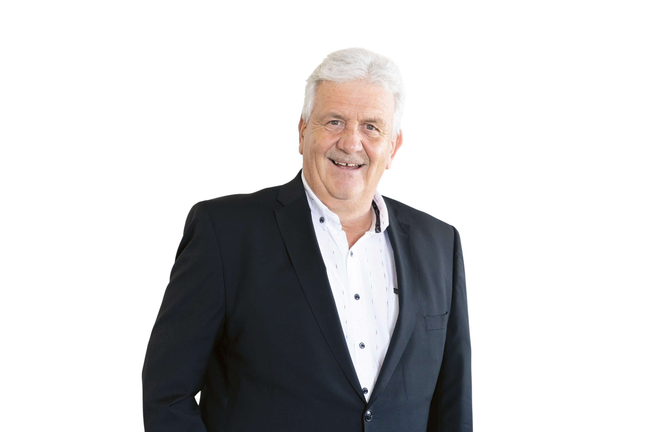 Ein älterer Mann mit weißem Haar, der eine dunkle Anzugsjacke und ein weißes Hemd mit Knöpfen trägt, lächelt, während er vor einem schlichten weißen Hintergrund steht.