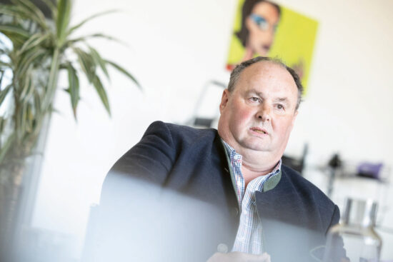 Ein Mann mittleren Alters in einem dunklen Blazer und einem karierten Hemd sitzt in einem Haus und schaut leicht zur Seite. Links steht eine verschwommene Grünpflanze und hinter ihm sind bunte Kunstwerke an der Wand zu sehen. Das Bild ist hell beleuchtet.