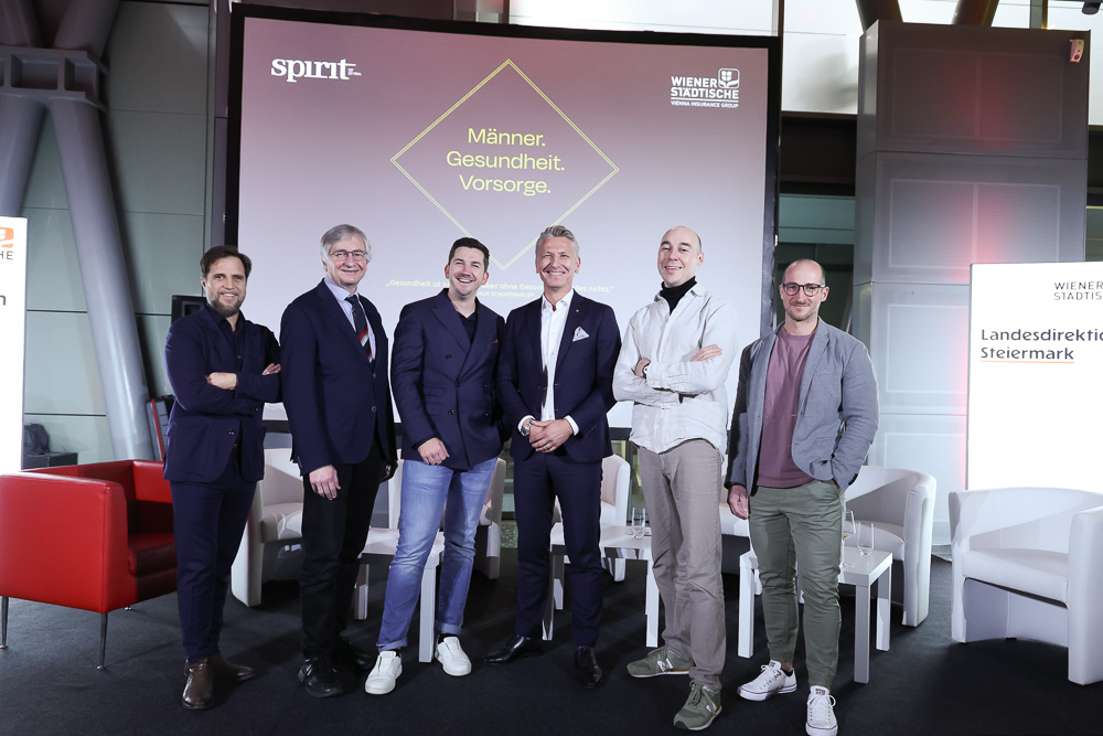 Sechs Männer stehen lächelnd vor einem Bildschirm mit der Aufschrift "Männer. Gesundheit. Vorsorge." bei einer feierlichen Veranstaltung und unterstreichen damit ihr Engagement für Initiativen wie die Ortskernbelebung Steiermark. Gekleidet sind sie in Smart-Casual- bis Business-Kleidung.