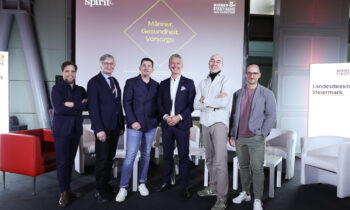 Sechs Männer in Business- und eleganter Freizeitkleidung stehen lächelnd auf der Bühne des Abends der Männergesundheit, hinter ihnen eine Leinwand zum Thema Männergesundheit und Prävention.