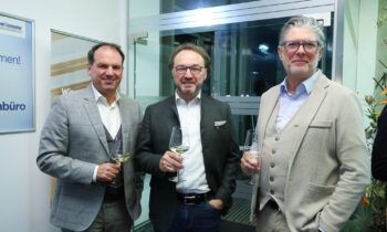 Drei Männer in Anzügen stehen bei der Veranstaltung "Abend der Männergesundheit" in einem Innenraum, jeder hält ein Glas Weißwein in der Hand und lächelt in die Kamera.