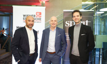 Drei Männer in Business-Kleidung lächeln drinnen zwischen zwei Markenbannern beim Abend der Männergesundheit für die Wiener Städtische und das Spirit Magazin.