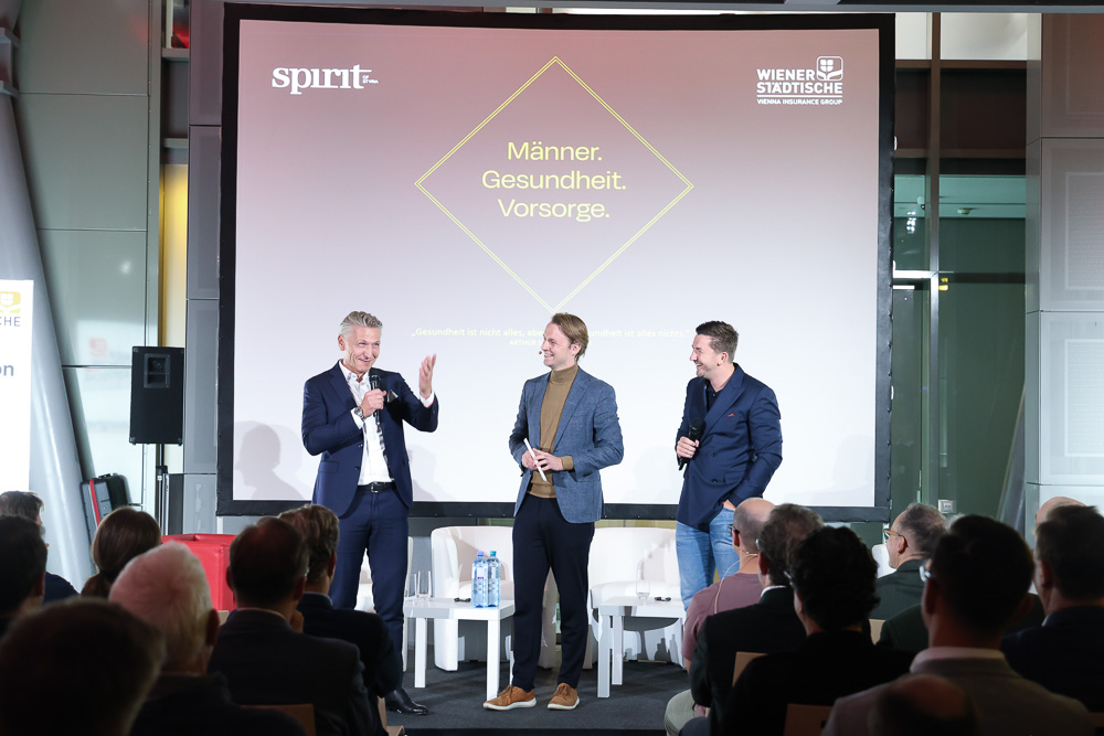 Drei Männer in Anzügen stehen auf der Bühne und sprechen und lächeln vor einem Publikum. Hinter ihnen wird auf einer Leinwand "Männer. Gesundheit. Vorsorge." bei einer Gesundheitsveranstaltung zum Thema Ortskernbelebung Steiermark.