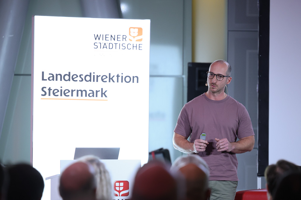Ein Mann in einem rosafarbenen T-Shirt spricht vor einem Publikum, das neben einem leuchtenden Schild mit der Aufschrift "WIENER STÄDTISCHE Landesdirektion Steiermark" steht, das auf die Bemühungen um die Ortskernbelebung in der Steiermark hinweist.