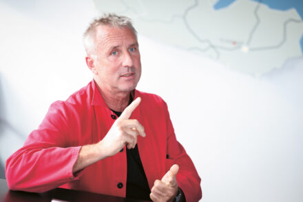 Ein Mann mit kurzen grauen Haaren und roter Jacke sitzt an einem Tisch und hebt einen Finger, als wolle er auf etwas hinweisen, während im Hintergrund eine Landkarte zu sehen ist.