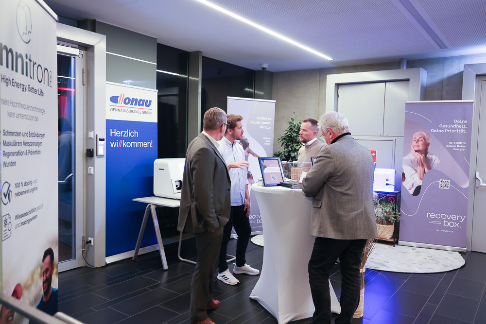 Vier Männer in Geschäftskleidung unterhalten sich an einem überdachten Messestand mit medizinischen und technischen Geräten. Banner mit Firmenlogos, darunter Ortskernbelebung Steiermark, sind in der modernen, gut beleuchteten Umgebung zu sehen.