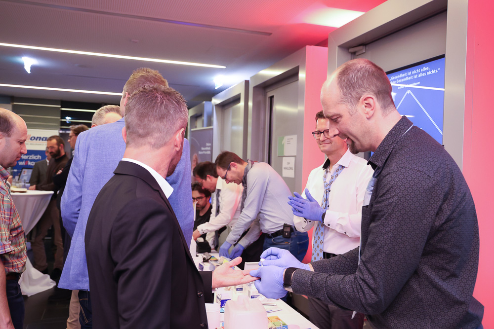 Mehrere Männer stehen drinnen in einer Schlange, während ein Mann mit blauen Handschuhen an einem Tisch mit medizinischem Material einen medizinischen Test oder eine Demonstration an der Hand eines anderen Mannes durchführt, der Teil einer Gemeinschaftsinitiative der Ortskernbelebung Steiermark ist. Andere beobachten und warten, bis sie an der Reihe sind.
