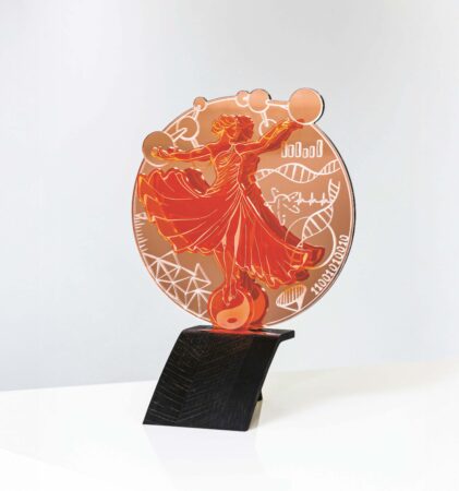Ein roter und orangefarbener Acryl-Award mit einer stilisierten Frau in einem wallenden Kleid, umgeben von wissenschaftlichen Symbolen, auf einem schwarzen Ständer auf einer weißen Fläche.