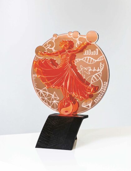 Ein roter und orangefarbener Acryl-Award mit einer stilisierten Frau in einem wallenden Kleid, umgeben von wissenschaftlichen Symbolen, auf einem schwarzen Ständer auf einer weißen Fläche.