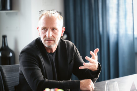 Ein Mann mit Brille auf dem Kopf gestikuliert mit der Hand, während er an einem Tisch sitzt und spricht, mit einem dunklen Vorhang im Hintergrund.