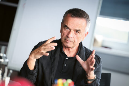 Ein Mann in einem schwarzen Hemd gestikuliert mit den Händen, während er spricht. Er sitzt in einem Innenraum, der Hintergrund ist unscharf und hinter ihm befindet sich ein Whiteboard.