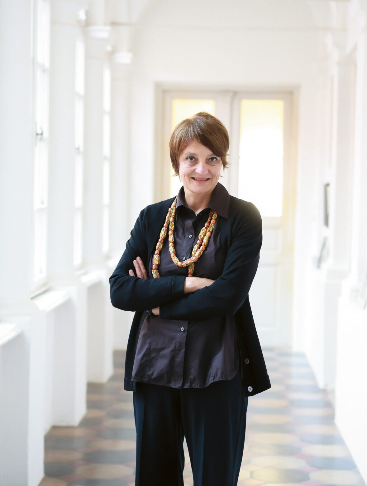Eine Frau mit kurzen Haaren steht mit verschränkten Armen in einem hellen Korridor, der von Fenstern und Kunstwerken an den Wänden gesäumt ist.