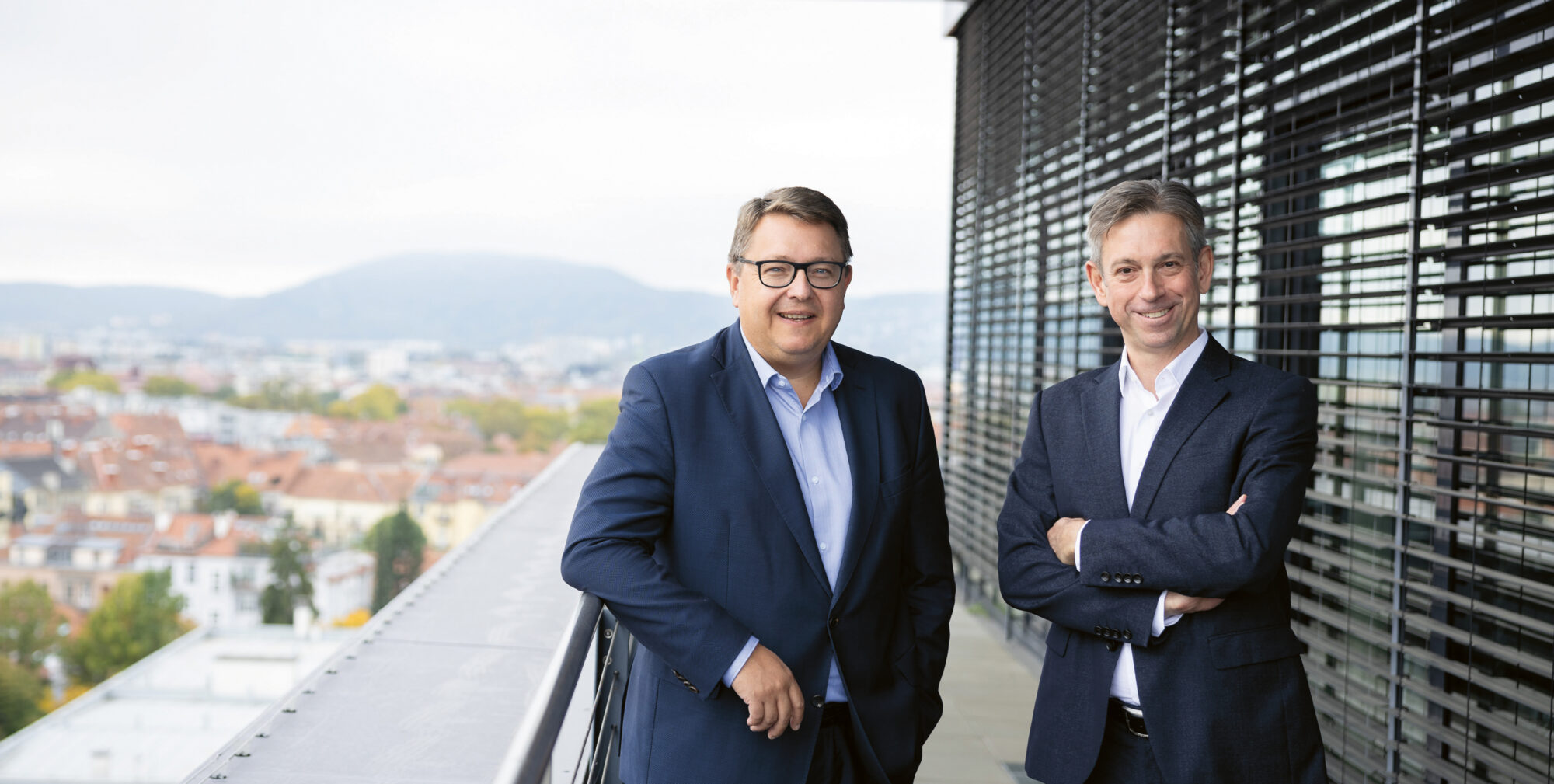 Zwei Männer in Anzügen stehen auf einem modernen Balkon mit einer Stadtlandschaft und Hügeln im Hintergrund. Ein Mann steht mit voreinander verschränkten Händen, der andere mit verschränkten Armen. Beide lächeln und wirken professionell.