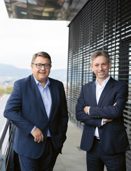 Zwei Männer in Anzügen stehen auf einem modernen Balkon mit einer Stadtlandschaft und Hügeln im Hintergrund. Ein Mann steht mit voreinander verschränkten Händen, der andere mit verschränkten Armen. Beide lächeln und wirken professionell.