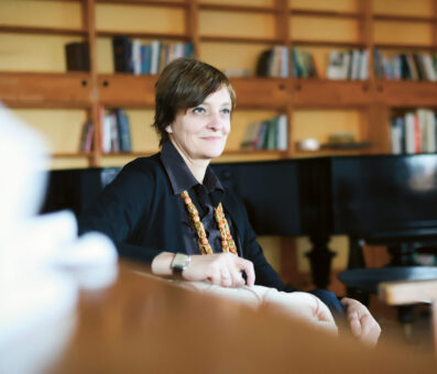 Eine Frau sitzt in einem gemütlichen Raum mit Bücherregalen und einem Klavier im Hintergrund und wirkt nachdenklich und entspannt.