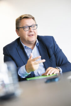 Ein Mann im blauen Anzug und mit Brille sitzt an einem Tisch und gestikuliert mit seiner Hand, während er spricht. Der Hintergrund ist einfarbig und unscharf, und auf dem Tisch sind ein grüner Ordner und ein Glas zu sehen.