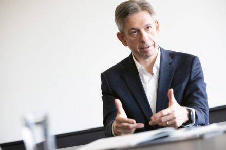 Ein Mann in einem dunklen Anzug und weißem Hemd gestikuliert mit den Händen, während er an einem Tisch spricht, mit einem verschwommenen Glas Wasser und Dokumenten im Vordergrund. Der Hintergrund ist schlicht und gut beleuchtet.