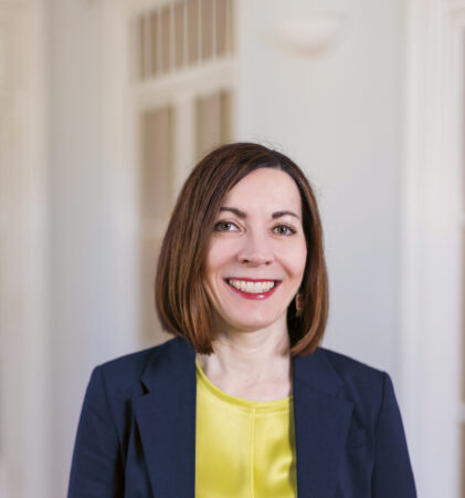 Eine Frau mit glattem braunem Haar, die einen marineblauen Blazer und ein gelbes Oberteil trägt, lächelt, während sie in einem hellen Raum mit Holzfußboden und Glastüren steht.