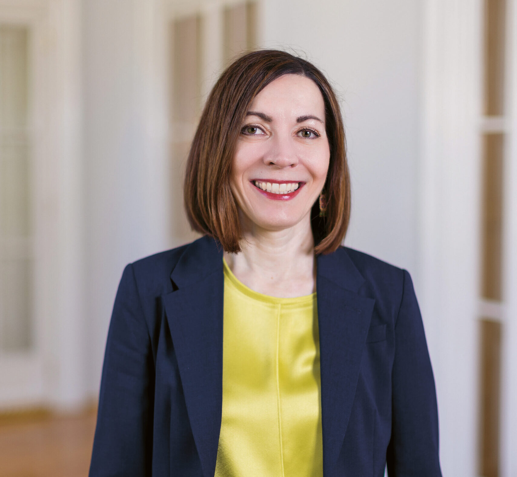 Eine Frau mit glattem braunem Haar, die einen marineblauen Blazer und ein gelbes Oberteil trägt, lächelt, während sie in einem hellen Raum mit Holzfußboden und Glastüren steht.