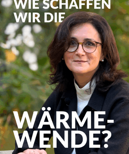 Eine Frau mit dunklem Haar und Brille sitzt an einem Tisch im Freien und lächelt leicht. Sie trägt eine schwarze Jacke und einen weißen Rollkragenpullover. Der Text auf Deutsch lautet: "Wie schaffen wir die Wärmewende?"