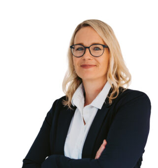 Eine selbstbewusste Frau mit blondem Haar und Brille, die einen dunklen Blazer und ein weißes Hemd trägt, steht mit verschränkten Armen vor einem weißen Hintergrund.