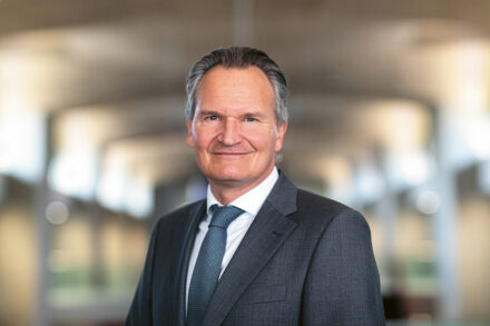 Ein Mann mittleren Alters in grauem Anzug und Krawatte lächelt, während er in einem Innenraum vor einem sanft verschwommenen Hintergrund aus Lichtern und Fenstern steht.