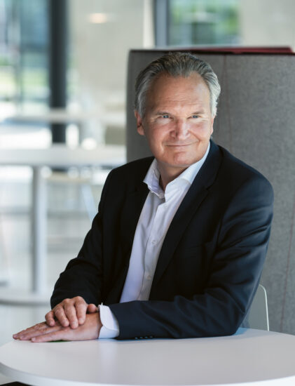 Ein Mann mittleren Alters in einem dunklen Anzug sitzt an einem runden weißen Tisch in einem modernen, hellen Büroraum, schaut zur Seite und lächelt leicht. Im Hintergrund sind leere weiße Stühle und Tische zu sehen.